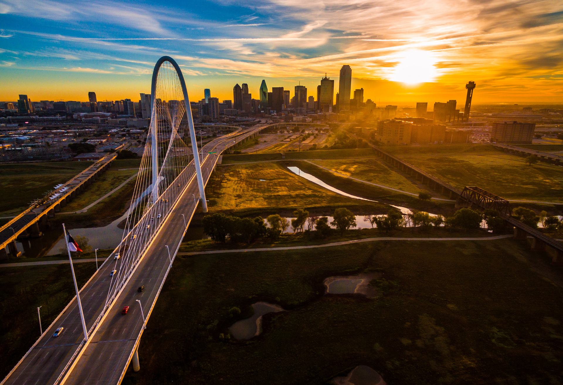 The downtown Dallas skyline at golden sunset beyond the arched Margaret Hunt Hill Bridge and the Trinity River greenbelt.