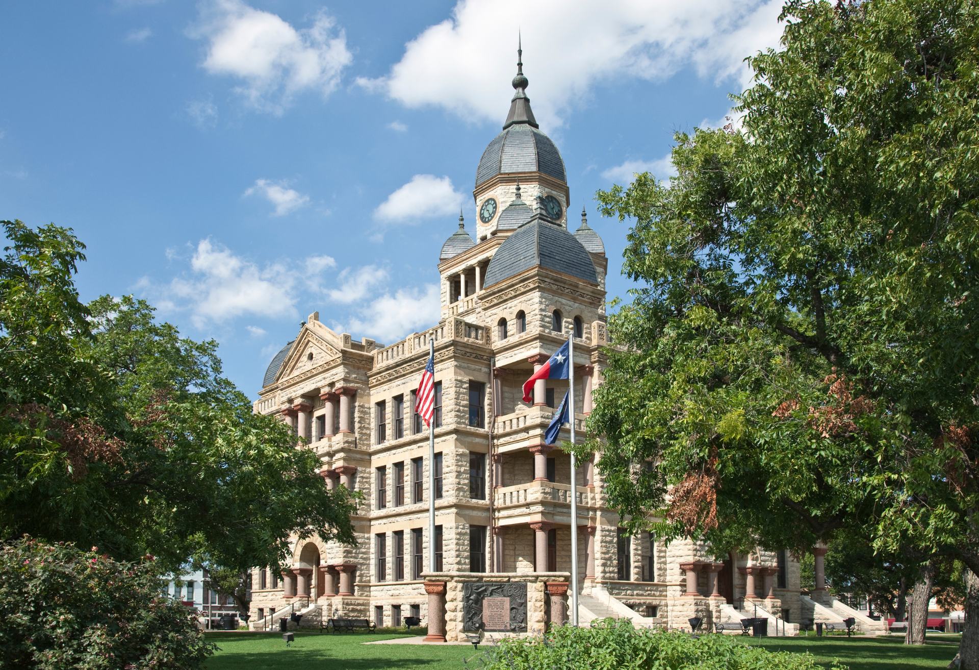 The historic Denton County Courthouse with its clock tower and copper domes, framed by trees on the downtown square.