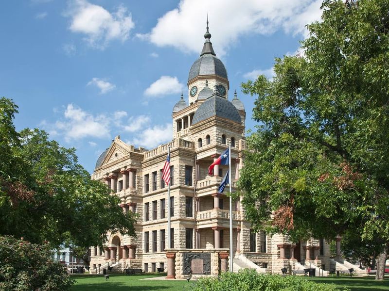 The historic Denton County Courthouse, a stone building with a domed clock tower, framed by mature green trees.