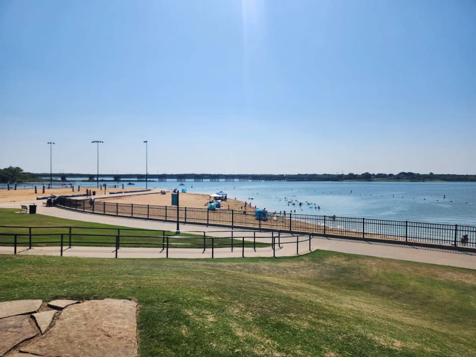 The sandy swimming beach and shoreline of Little Elm Park on Lewisville Lake, with a bridge in the distance.