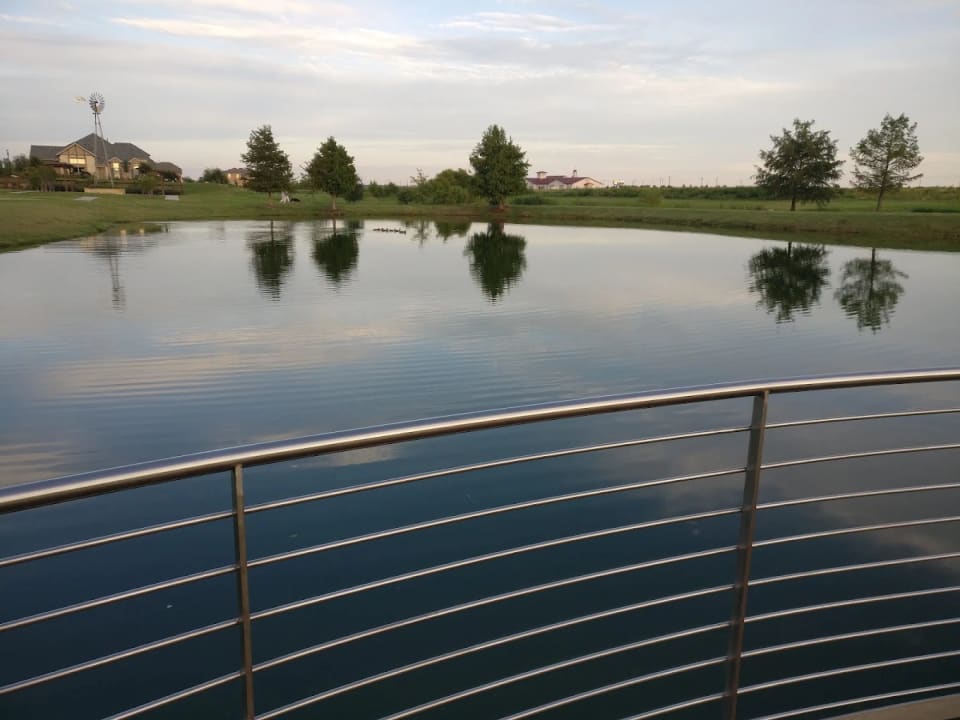 A community lagoon with a windmill, homes, and trees reflected in still water at Windsong Ranch in Prosper, Texas.