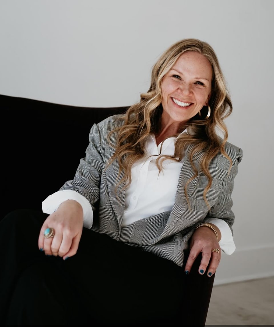 Realtor Sara Bigley, smiling in a houndstooth blazer and white blouse, seated in a black chair.
